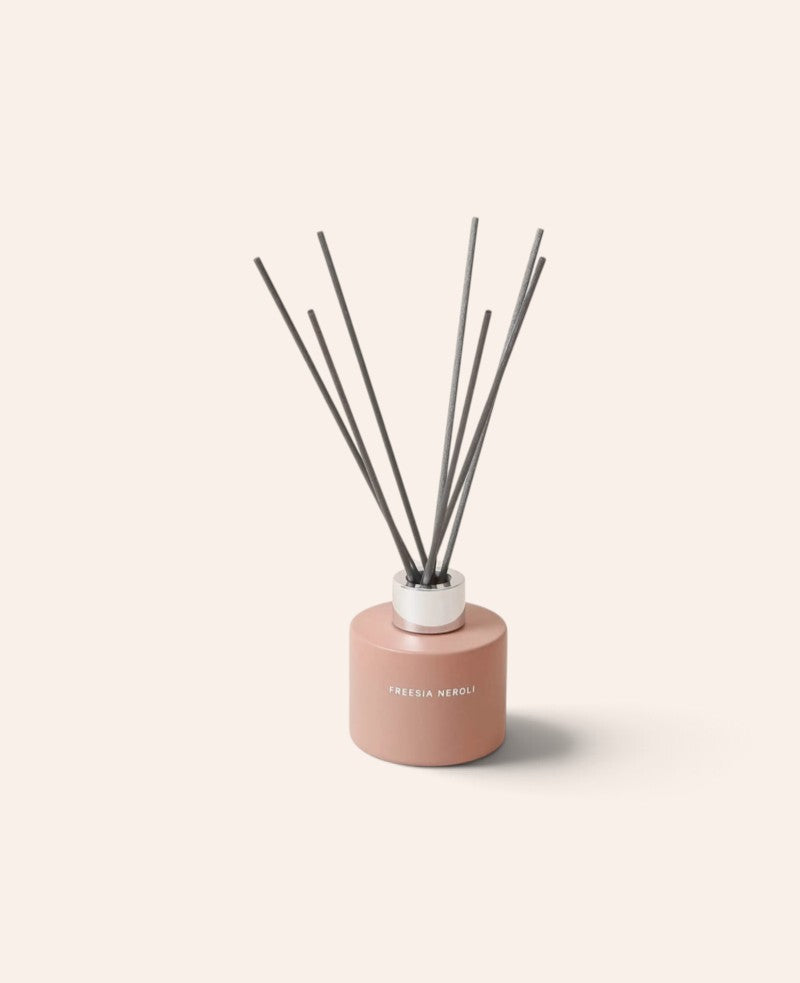 Esmée Duft Diffuser Freesia Neroli in einem matten, rosafarbenen Glasbehälter mit sieben grauen Holzfaser-Stäbchen.
