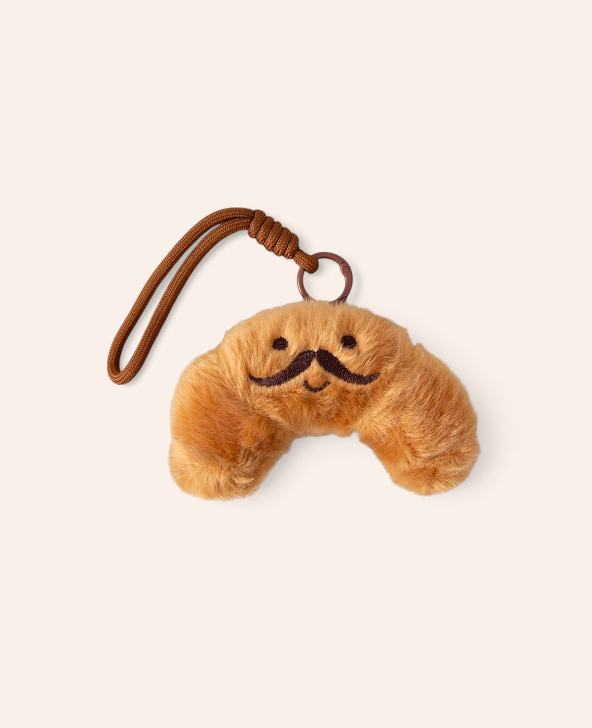 Esmée Taschenanhänger aus weichem, goldbraunem Plüsch in Croissant-Form mit aufgesticktem Schnurrbart und Schlaufe