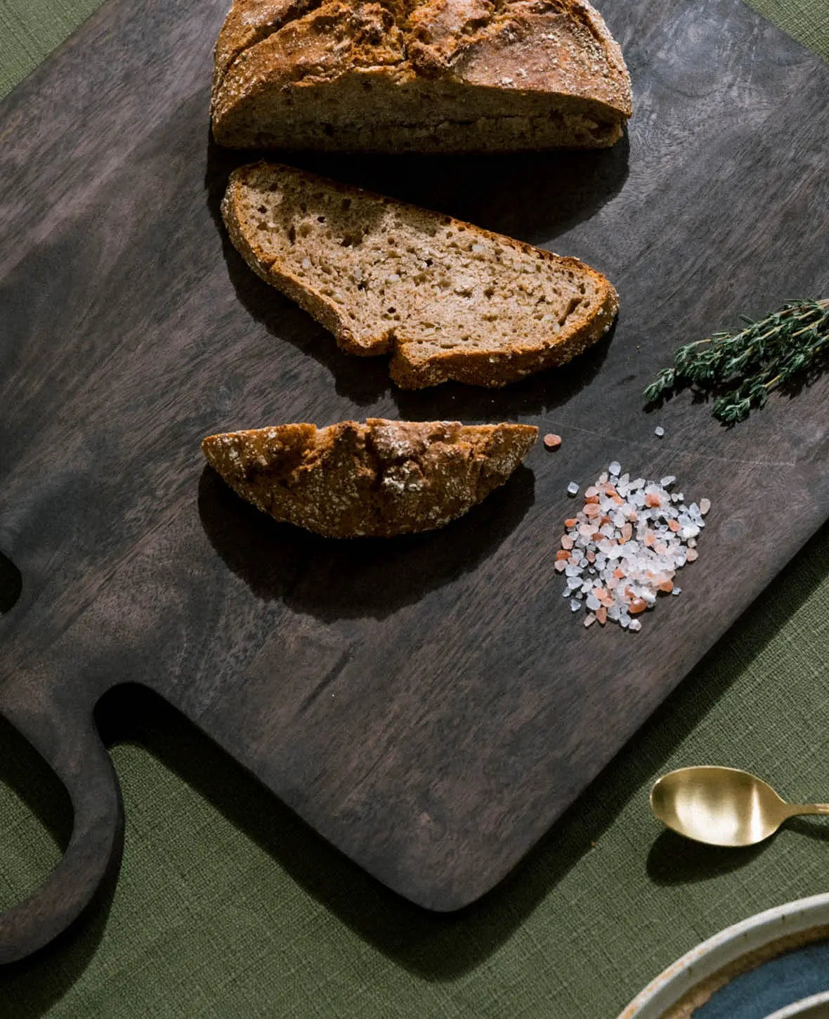 Esmée Schneidebrett aus dunkelbraunem Mangoholz mit Brot, Salz und Thymian auf grünem Untergrund
