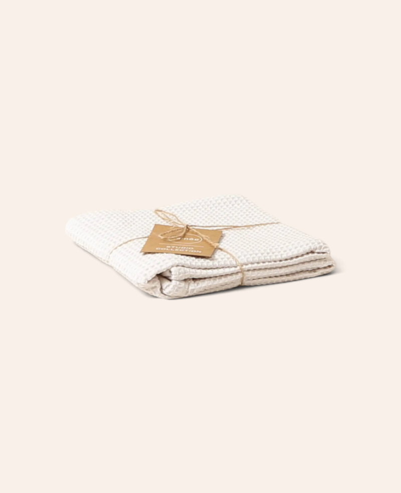 Esmée Handtuch aus off-white Baumwolle mit Waffel-Piqué-Struktur, gefaltet mit braunem Markenanhänger.